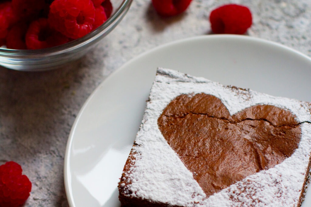 A brownie with a powdered sugar heart on a plate beside a bowl of raspberries