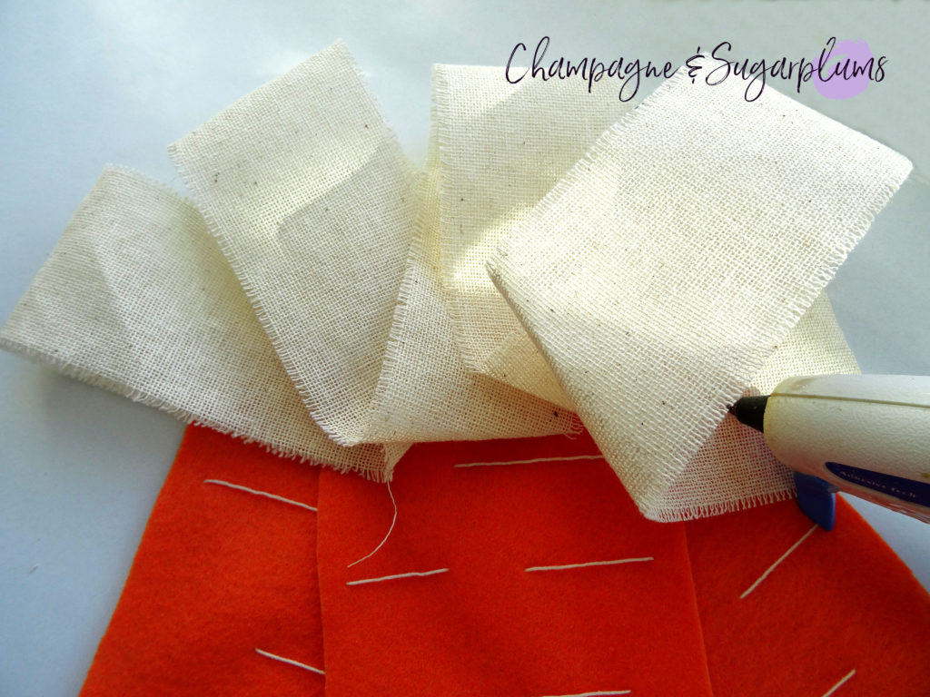 A glue gun being used to add thick cream ribbon to the top of three felt carrots by Champagne and Sugarplums