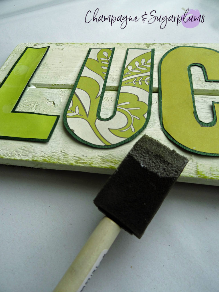 Decoupaging paper letters onto a wood plank with a foam brush by Champagne and Sugarplums