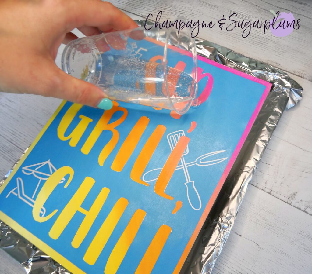 Pouring resin from a plastic cup over a wood sign on a tray covered with tin foil by Champagne and Sugarplums