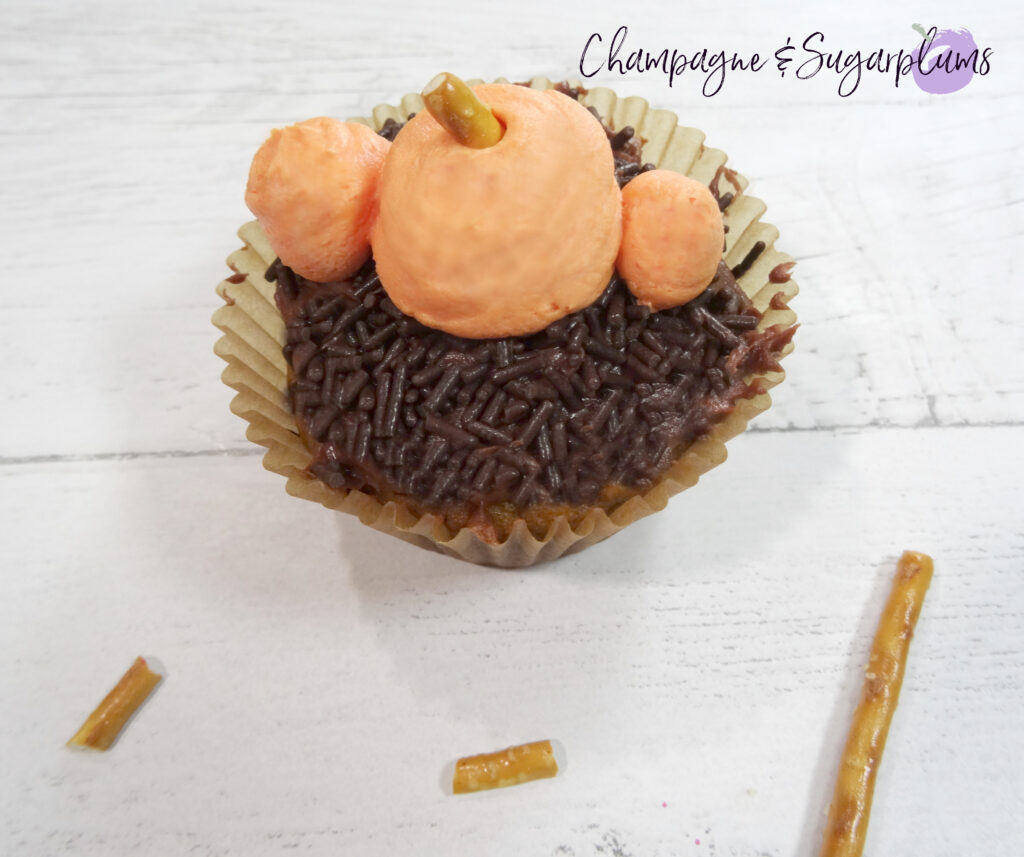 Adding pretzel stems to icing pumpkins on a chocolate cupcake on a white background by Champagne and Sugarplums