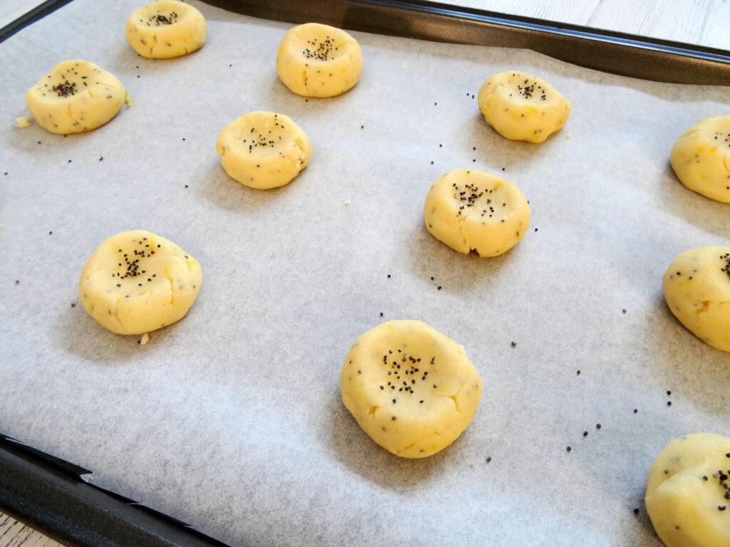 Cookie dough in small flattened balls, sprinkled with poppy seeds on a lined baking sheet by Champagne and Sugarplums