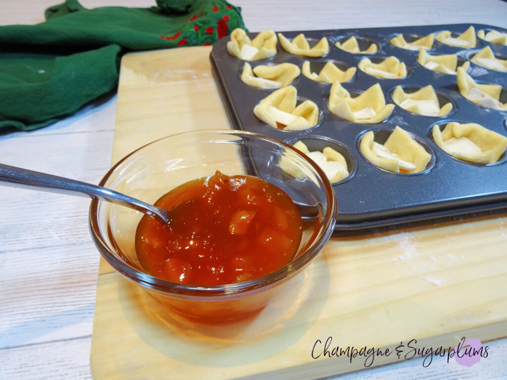 A bowl of mango chutney beside a mini muffin tin filled with dough squares with Brie pieces on a wood board by Champagne and Sugarplums