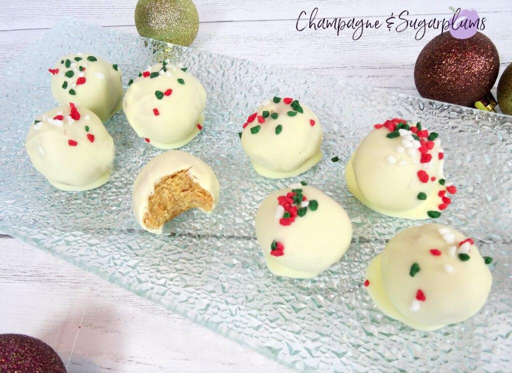 Natural Peanut Butter Snowballs, one with a bite in it, on a glass plate surrounded by ornaments on a white background by Champagne and Sugarplums 