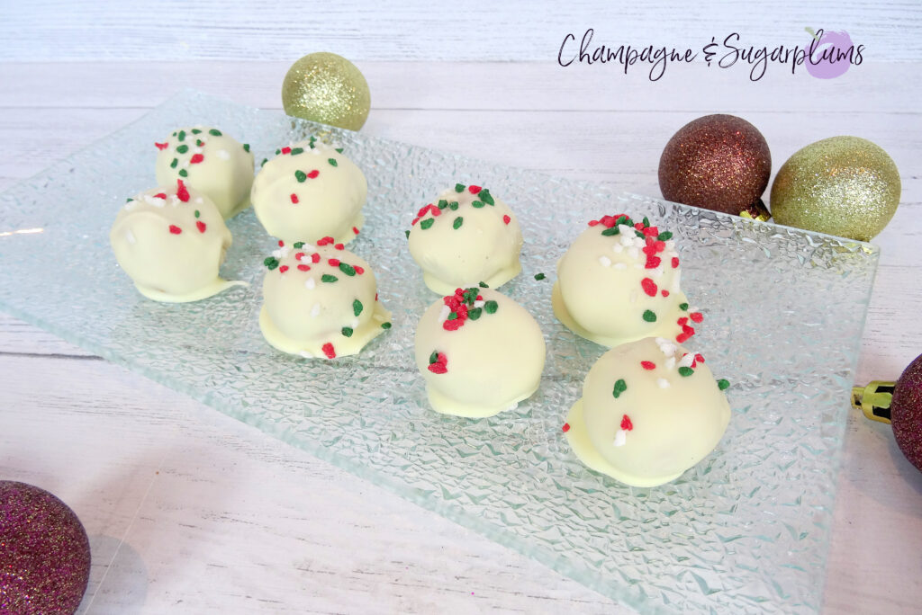 Natural Peanut Butter Snowballs on a glass plate surrounded by ornaments on a white background by Champagne and Sugarplums