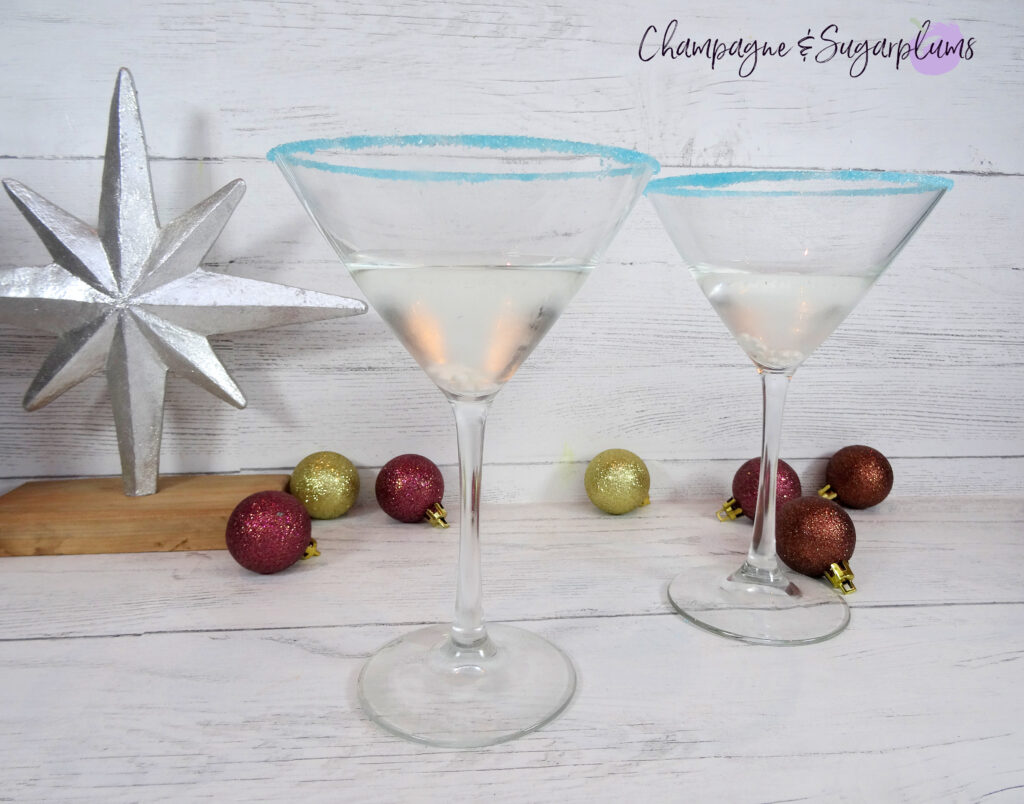 Polar Bear Martini cocktails, rimmed with blue sugar on a white background, surrounded by gold and brown mini ornaments by Champagne and Sugarplums