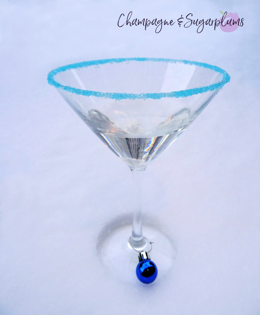 Polar Bear Martini cocktail, rimmed with blue sugar and decorated with an ornament shaped glass marker on a white background by Champagne and Sugarplums