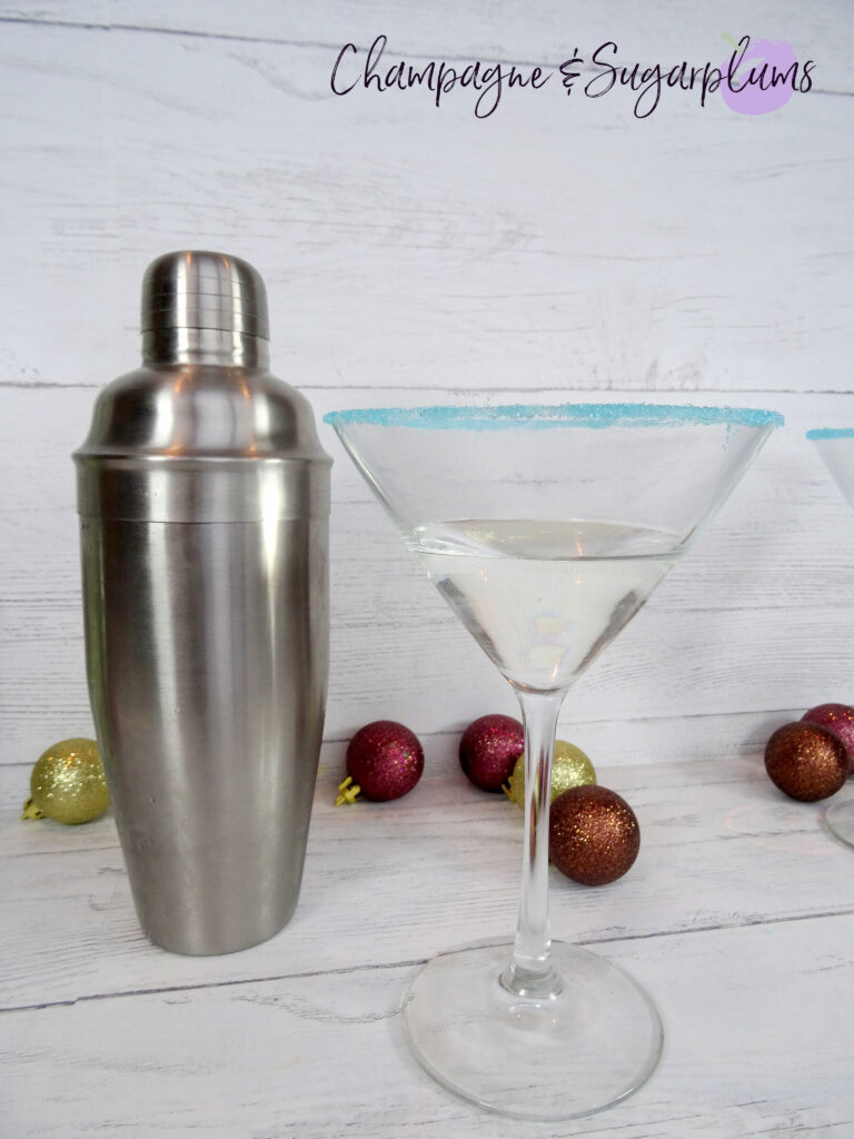 Polar Bear Martini cocktail, rimmed with blue sugar, sitting beside a cocktail shaker on a white background by Champagne and Sugarplums