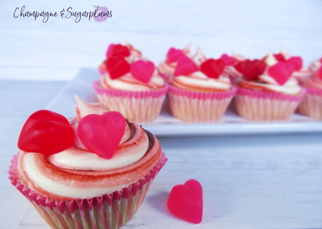 A Cherry Cupcake on a white background with a dozen more cupcakes on a white plate in the background by Champagne and Sugarplums