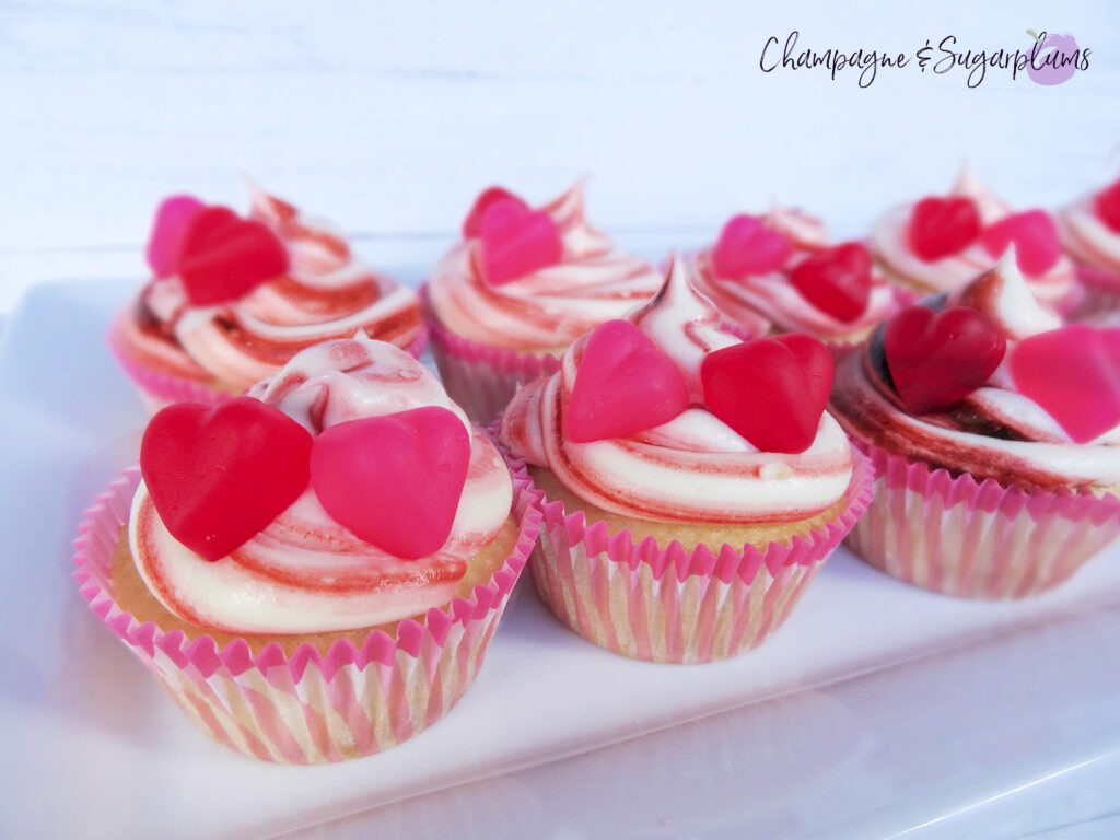 Cupcakes on a white plate decorated with pink striped icing and red and pink heart candies by Champagne and Sugarplums