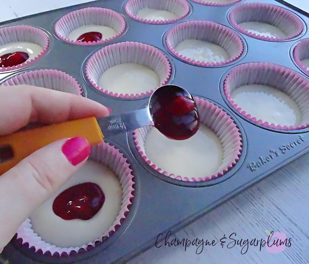 Adding cherry filling to vanilla cupcake mix in liners set inside of muffin tin by Champagne and Sugarplums