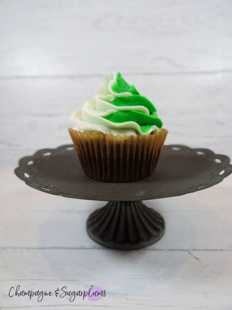 A green and white frosted cupcake sitting on a dark metal cake stand on a white background by Champagne and Sugarplums