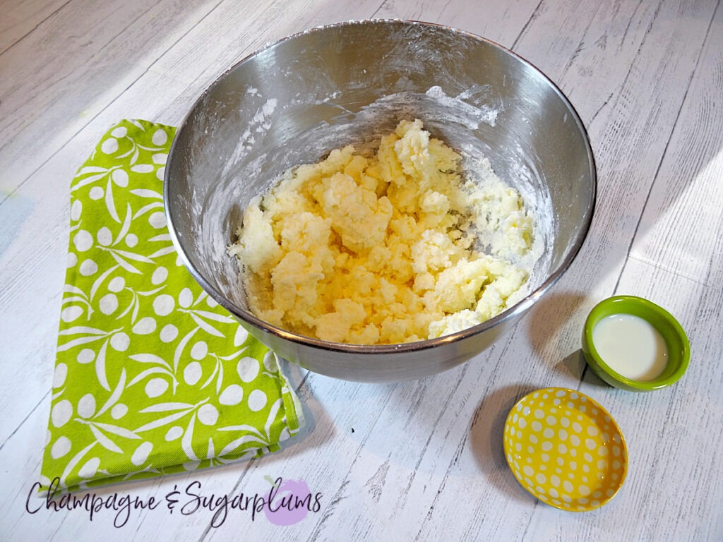 White frosting mixed in a metal mixing bowl beside a green tea cloth and green and yellow pinch bowls on a white background by Champagne and Sugarplums