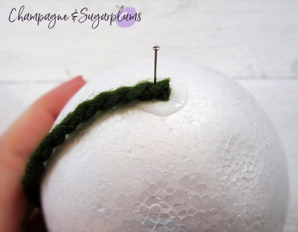 Sticking a pin into dark green yarn on a  Styrofoam ball on a white background by Champagne and Sugarplums