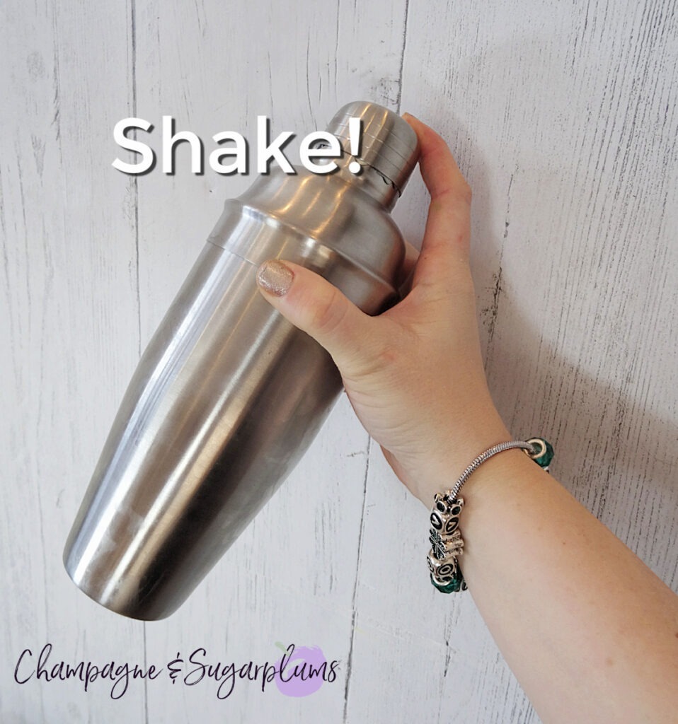 A metal cocktail shaker being held by a hand wearing a bracelet with the word 'shake' by Champagne and Sugarplums