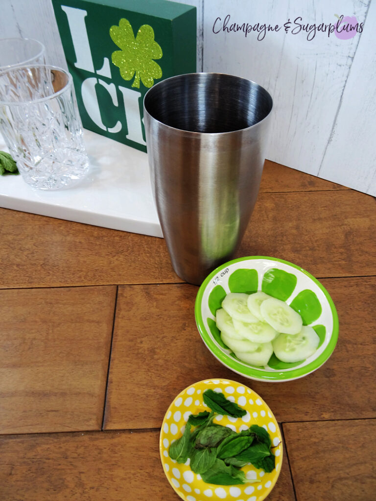 A metal cocktail shaker beside a bowl of cucumber slices and a bowl of mint leaves to make a cocktail by Champagne and Sugarplums