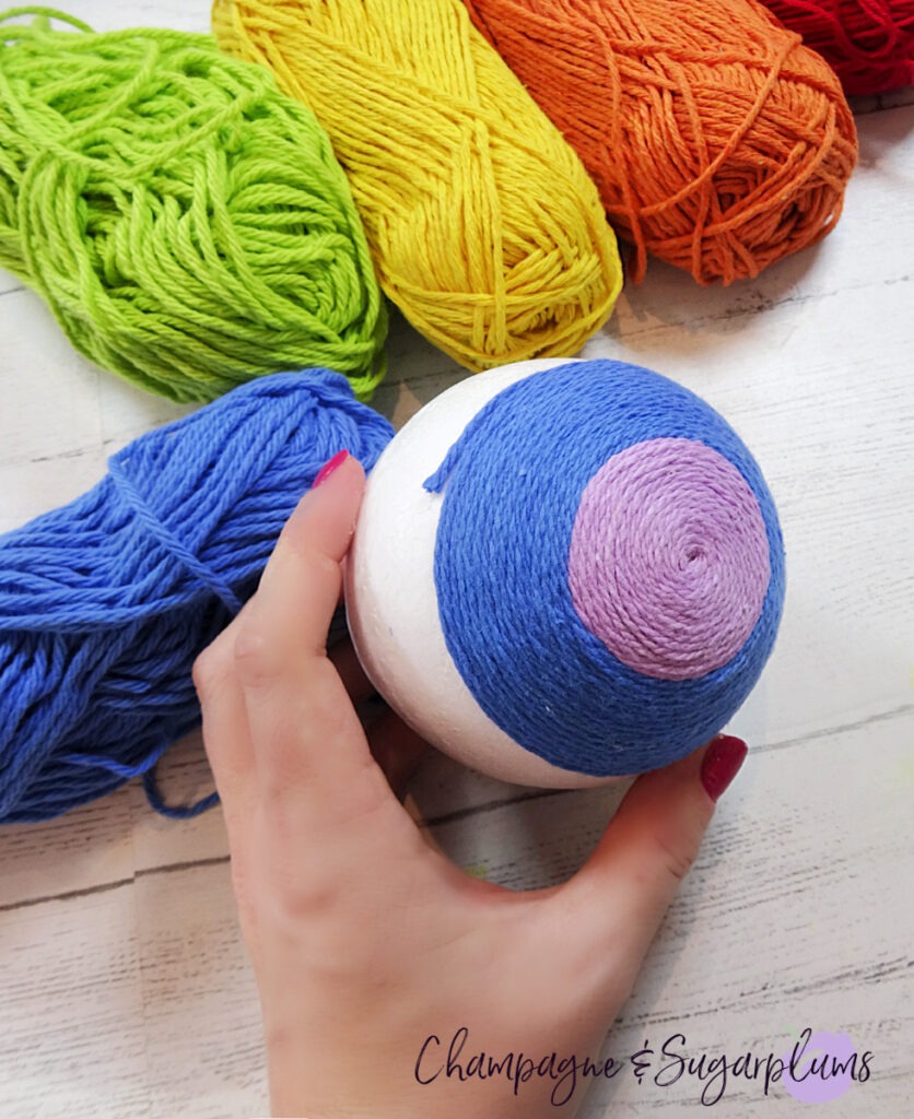 Wrapping blue yarn around a  Styrofoam ball to make a rainbow on a white background by Champagne and Sugarplums