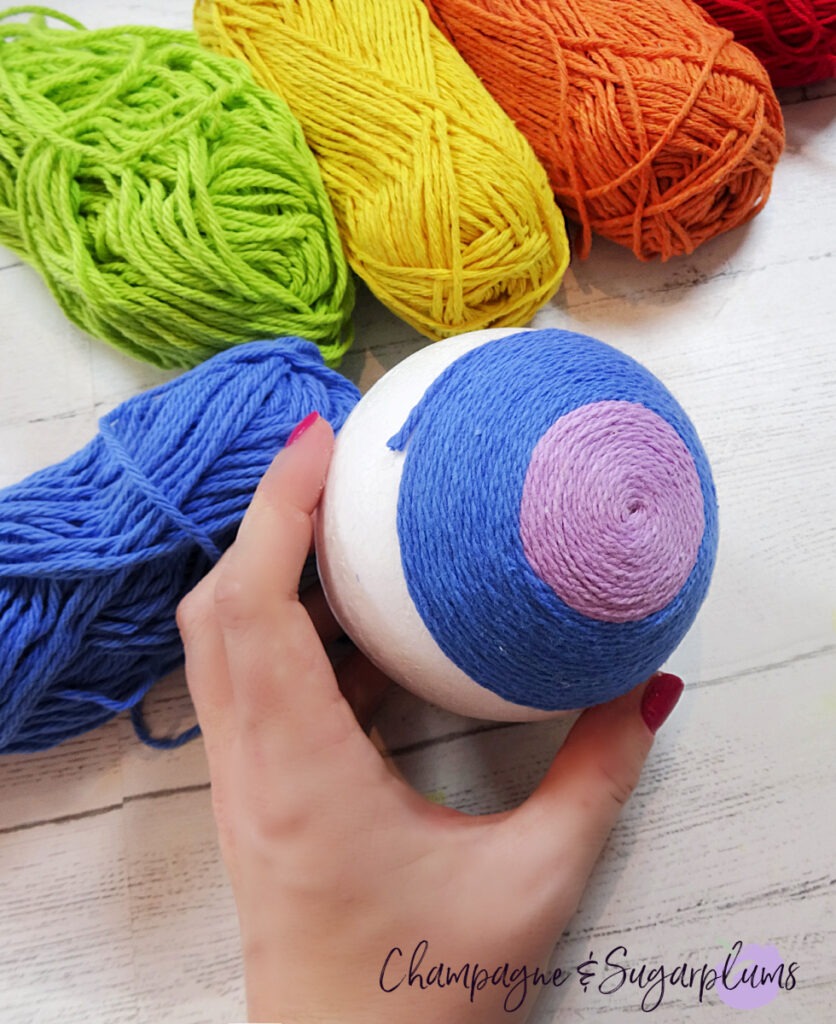 Wrapping blue yarn around a Styrofoam ball to make a rainbow on a white background by Champagne and Sugarplums
