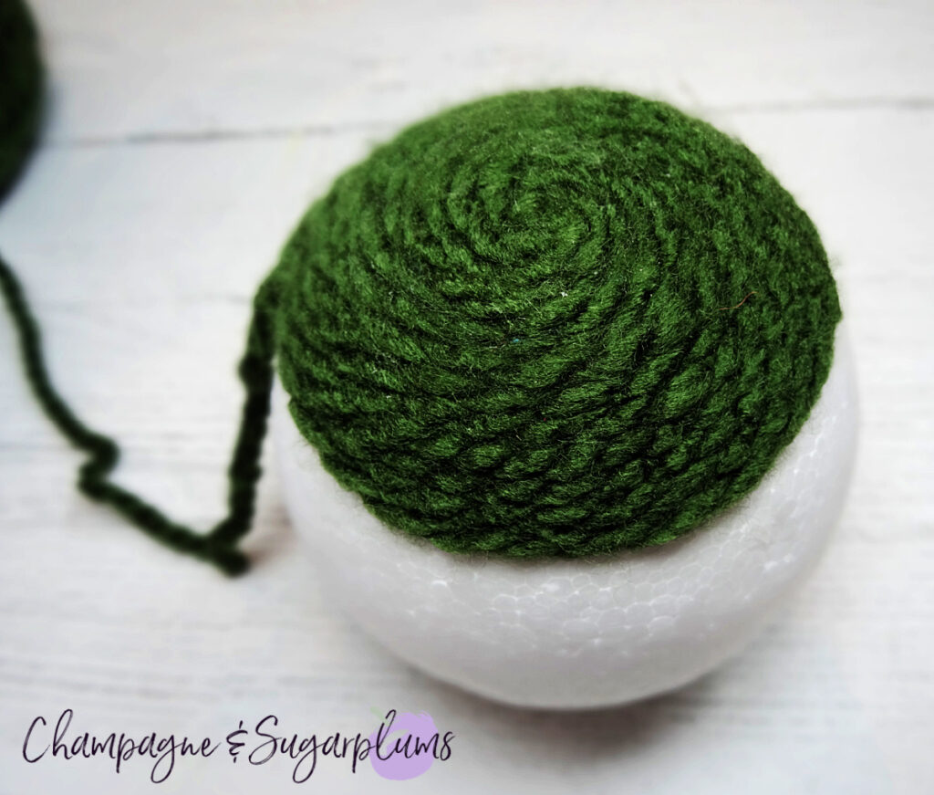 Wrapping dark green yarn around a Styrofoam ball on a white background by Champagne and Sugarplums