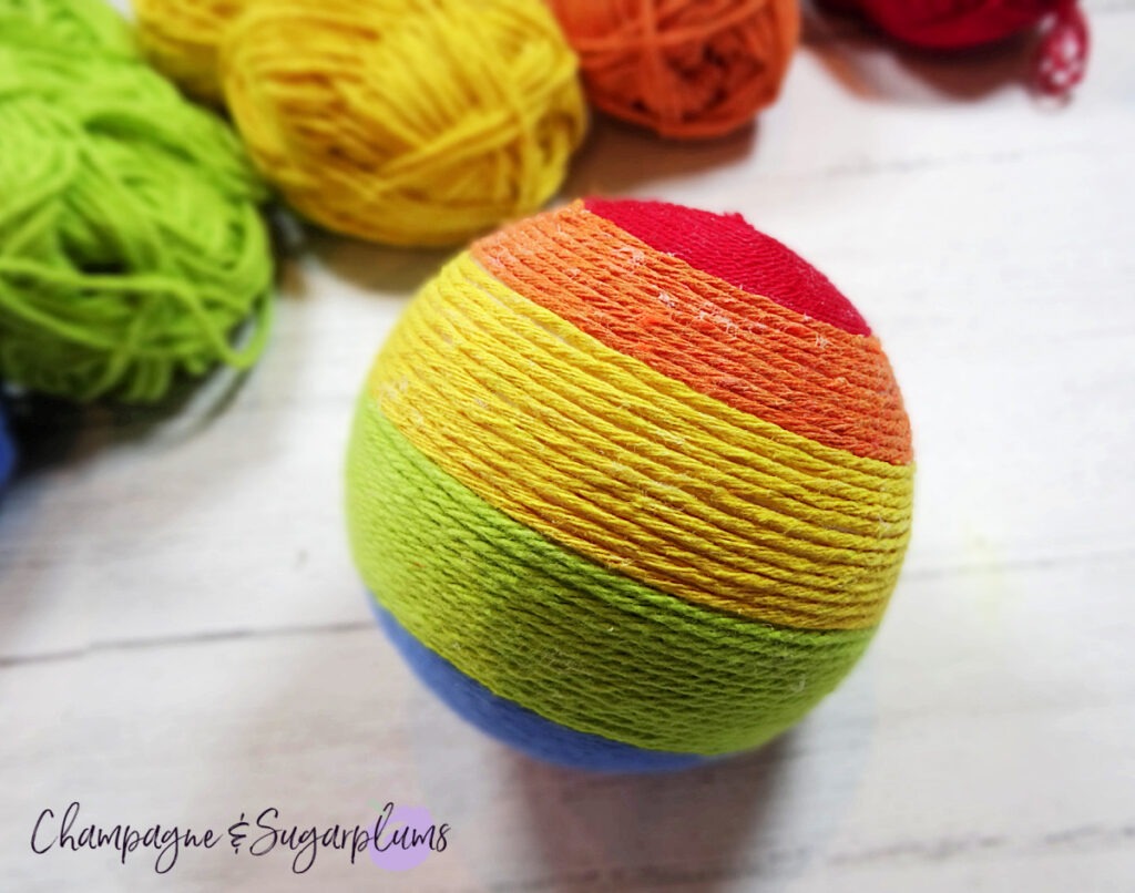 A Styrofoam ball wrapped in varying shades of yarn to make a rainbow on a white background by Champagne and Sugarplums