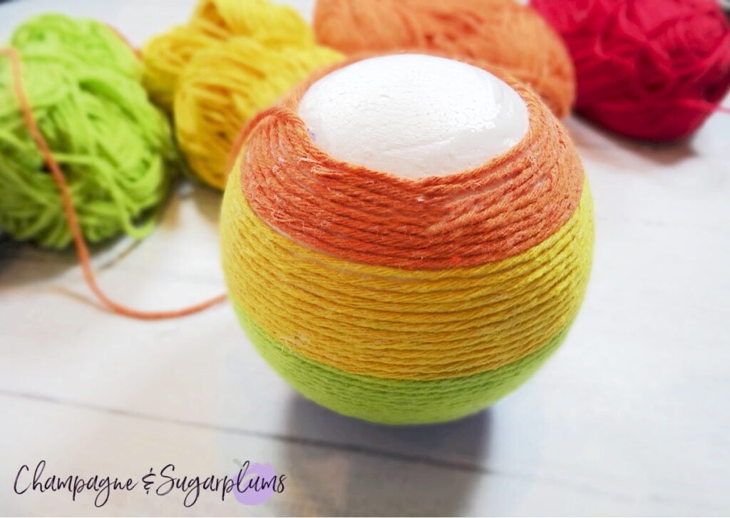 Wrapping orange yarn around a  Styrofoam ball to make a rainbow on a white background by Champagne and Sugarplums