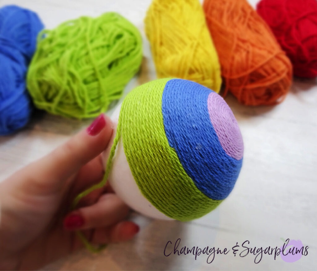 Wrapping green yarn around a Styrofoam ball to make a rainbow on a white background by Champagne and Sugarplums