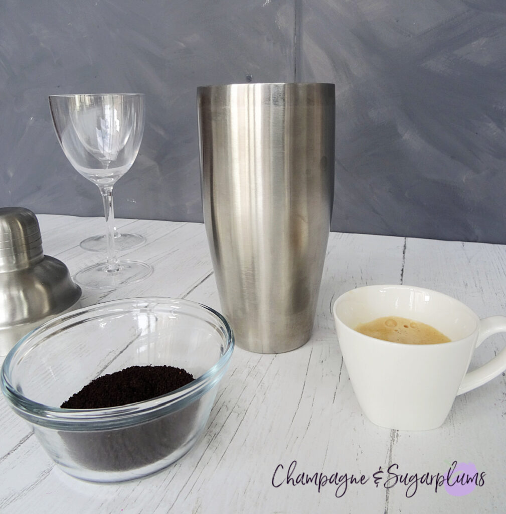 A steel cocktail shaker, with a shot of espresso and a bowl of coffee grounds on a white and grey background by Champagne and Sugarplums