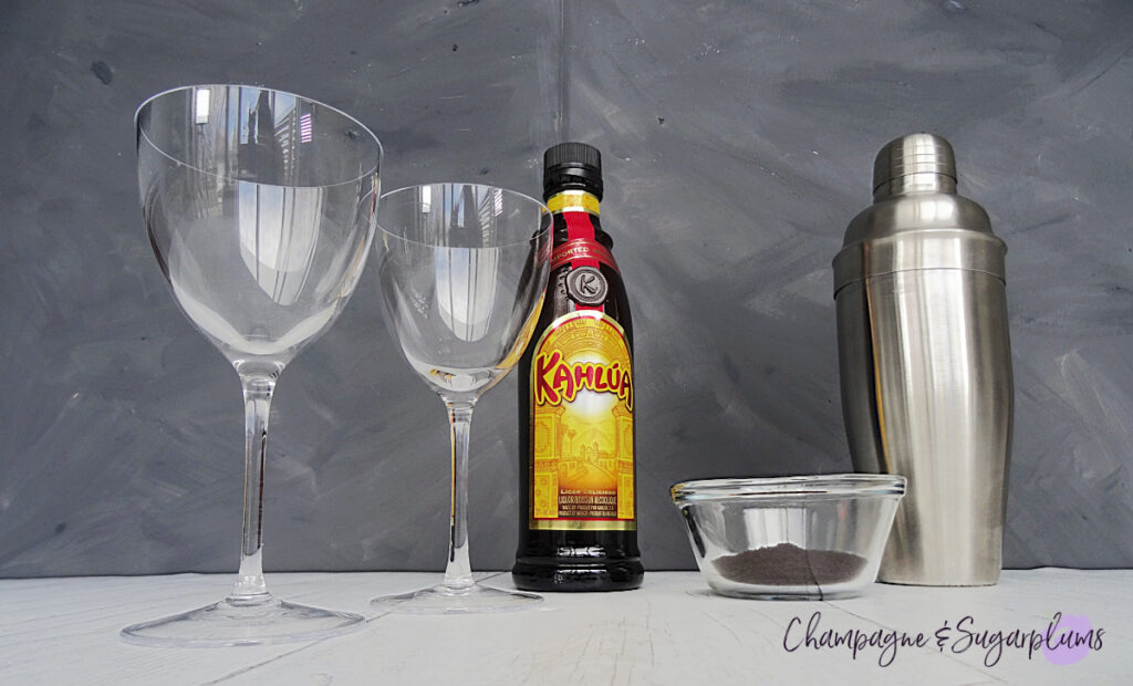 A steel cocktail shaker, with a bottle of Kahlua, Nick and Nora glasses and a bowl of coffee grounds on a white and grey background by Champagne and Sugarplums