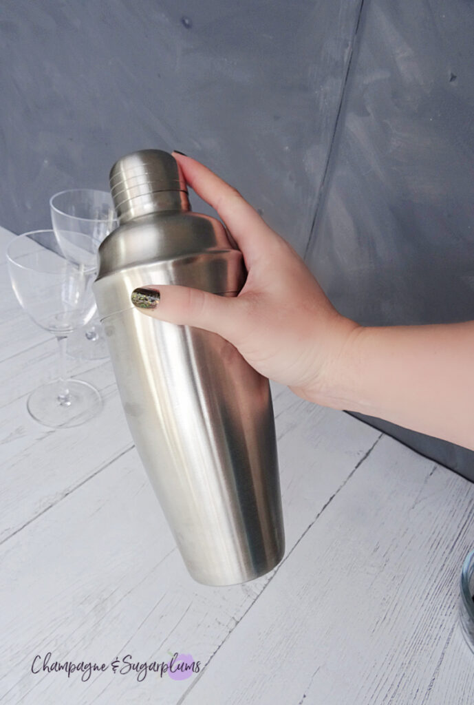 A hand shaking a steel cocktail shaker on a white and grey background by Champagne and Sugarplums
