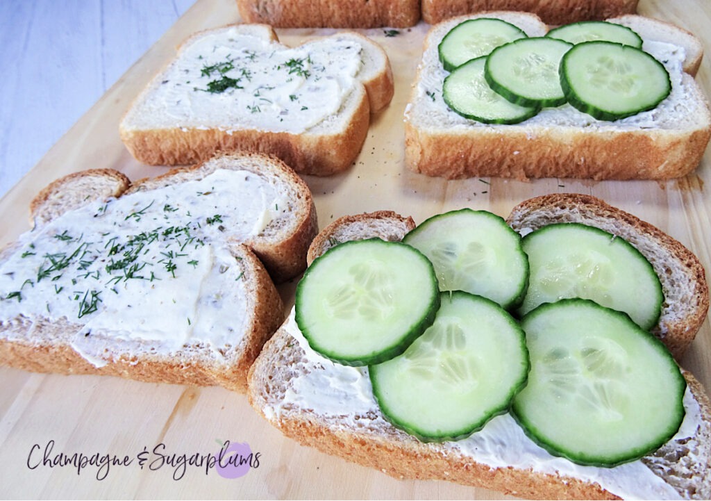 Assembling cucumber and cream cheese onto bread slices on a wood board by Champagne and Sugarplums