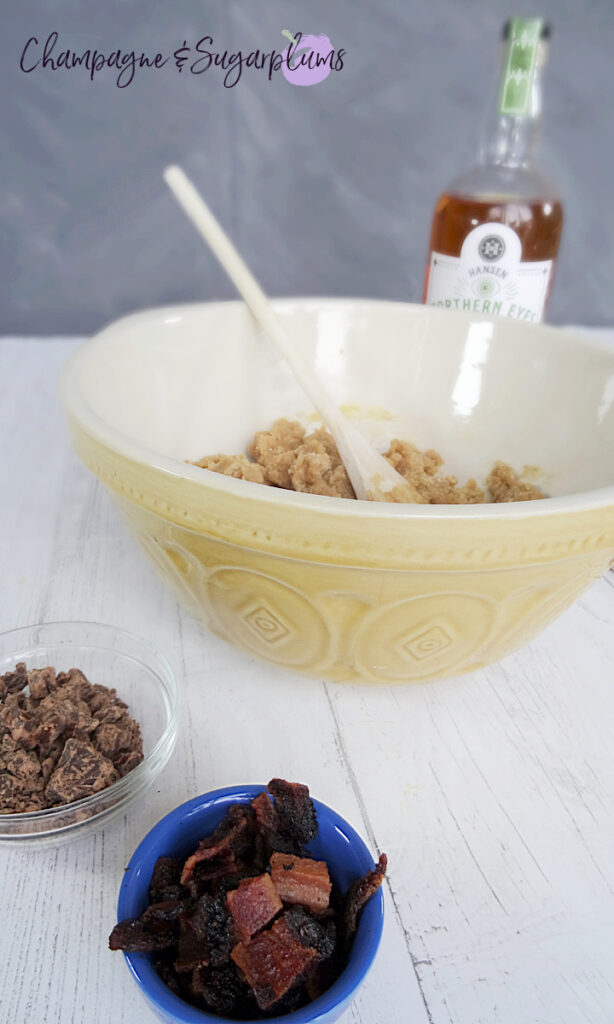 Cookie dough in a mixing bowl with a wooden spoon with a whiskey bottle, bowl of chocolate chunks and a bowl of bacon on a white background by Champagne and Sugarplums