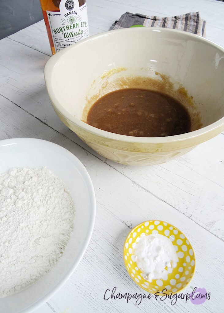 Cookie dough in a mixing bowl with a whiskey bottle, bowl of flour and a bowl of baking soda on a white background by Champagne and Sugarplums