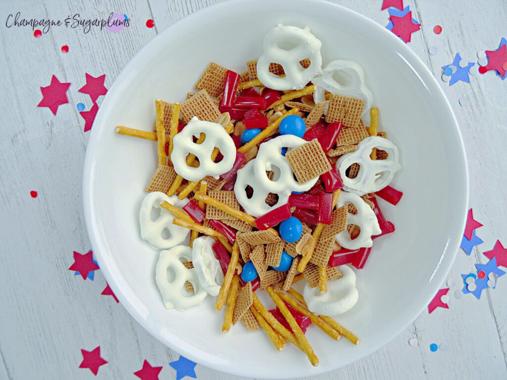 Fourth of July Snack Mix in a white bowl on a white background with blue and red stars and confetti by Champagne and Sugarplums