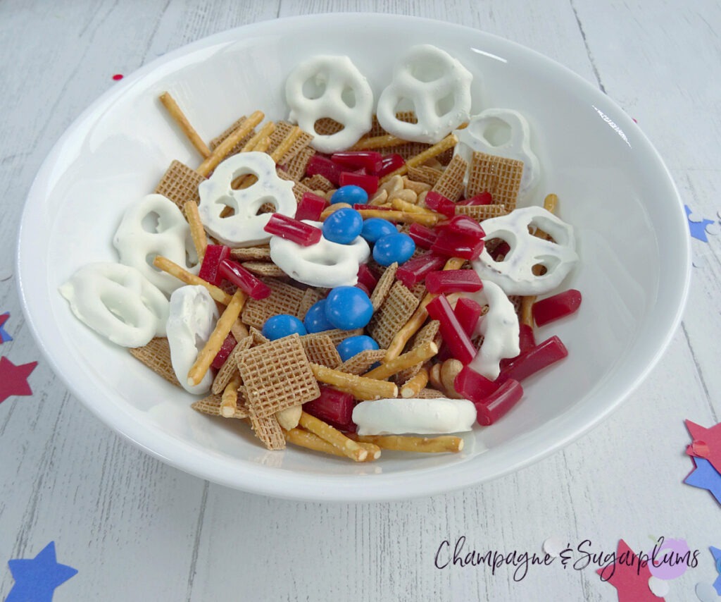 Snack Mix in a white bowl on a white background with blue and red stars and confetti by Champagne and Sugarplums