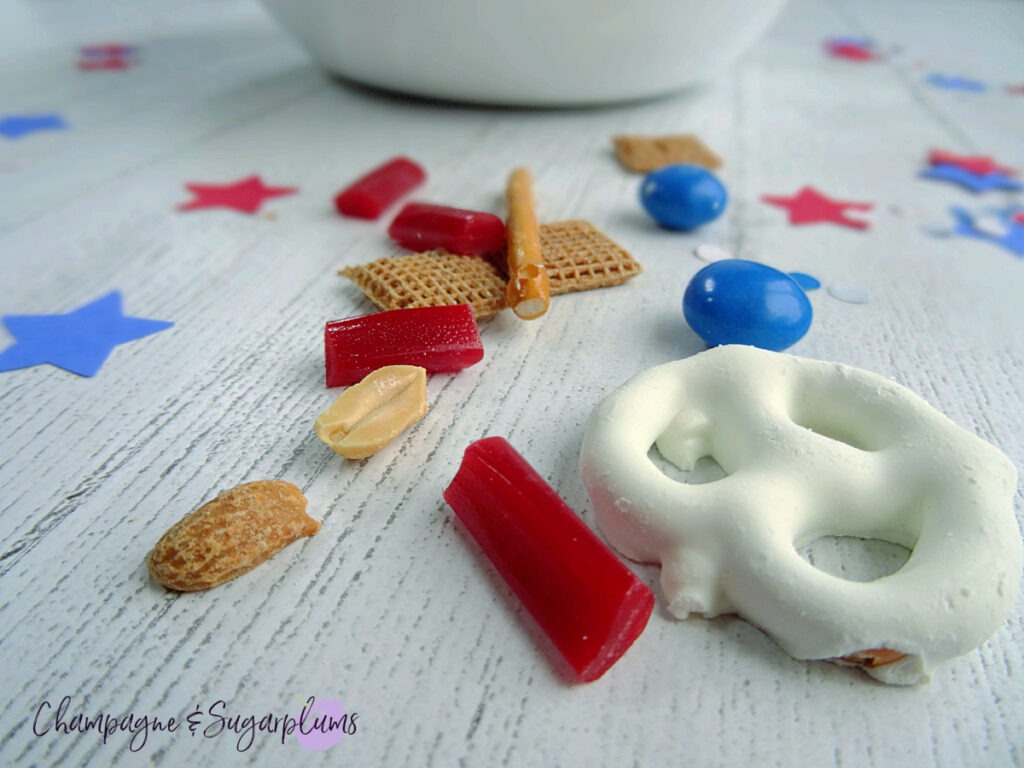 Fourth of July Snack Mix in a white bowl on a white background with blue and red stars and confetti by Champagne and Sugarplums