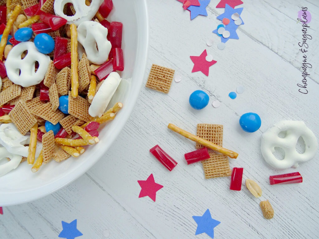 Fourth of July Snack Mix in a white bowl on a white background with blue and red stars and confetti by Champagne and Sugarplums