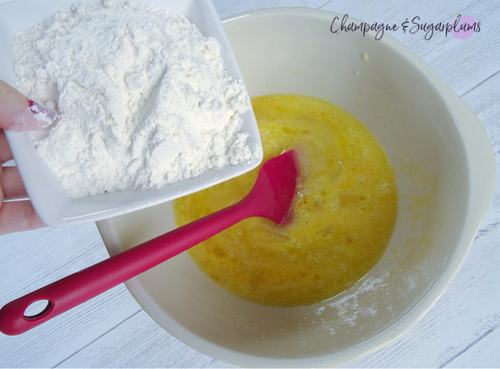 Adding flour to a mixing bowl with a red spatula and melted butter inside by Champagne and Sugarplums