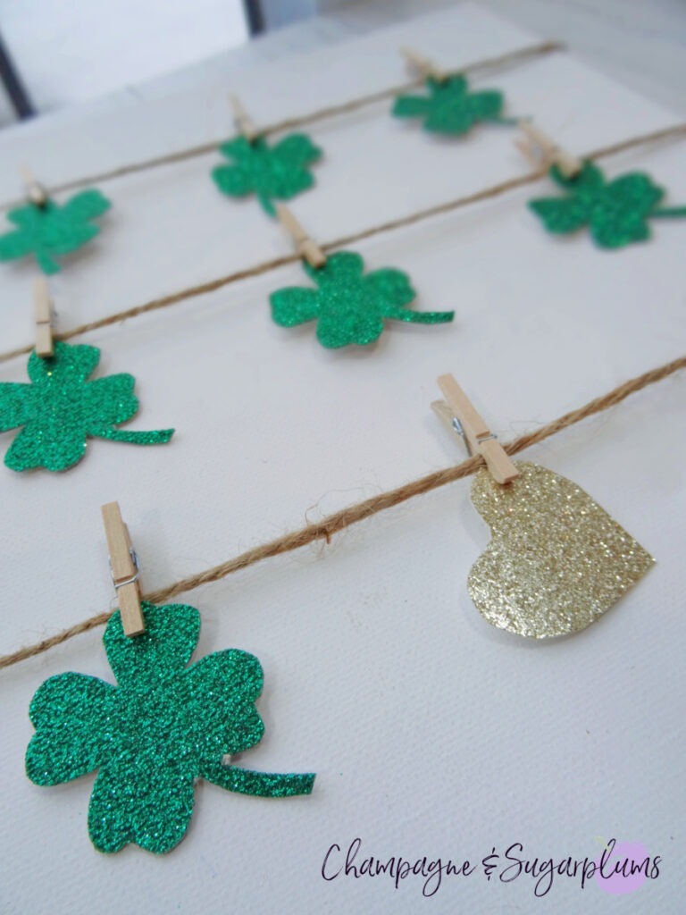 A close-up of a canvas, with green clover cut-outs hanging from tiny clothespins on twine by Champagne and Sugarplums