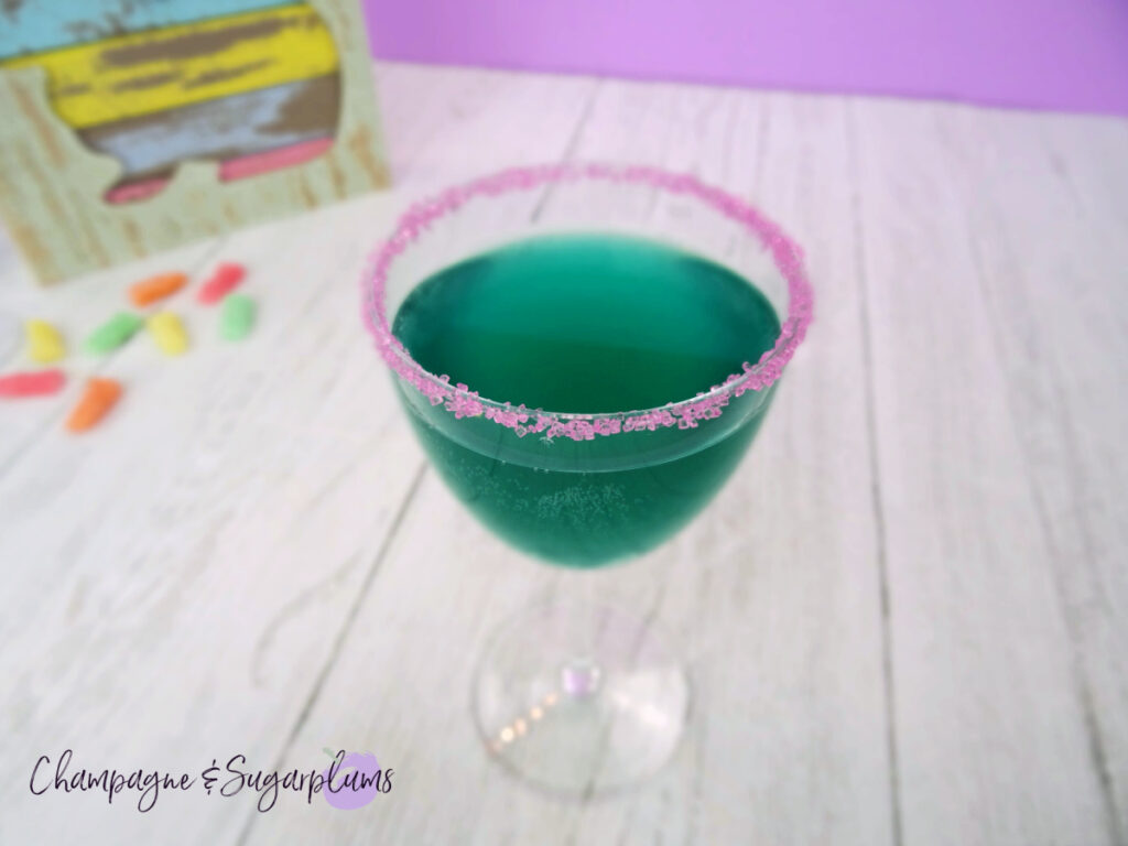 A teal cocktail with a pink sugared rim in a Nick and Nora glass on a white and purple background by Champagne and Sugarplums
