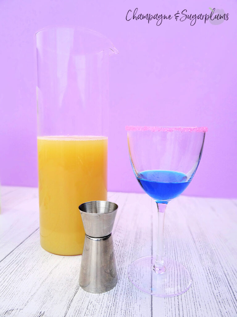 Mixing a cocktail, using blue curacao and pineapple juice in a Nick and Nora glass on a white and purple background by Champagne and Sugarplums