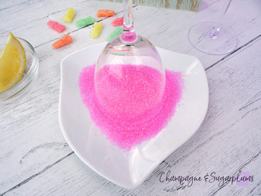 Sugaring the rim of a Nick and Nora glass, with a lemon slice and pink sugar on a white plate, on a white background by Champagne and Sugarplums