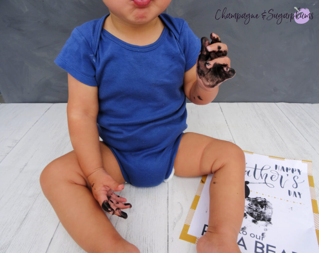 Papa Bear Handprint craft on a white background, with a toddler sitting beside it by Champagne and Sugarplums