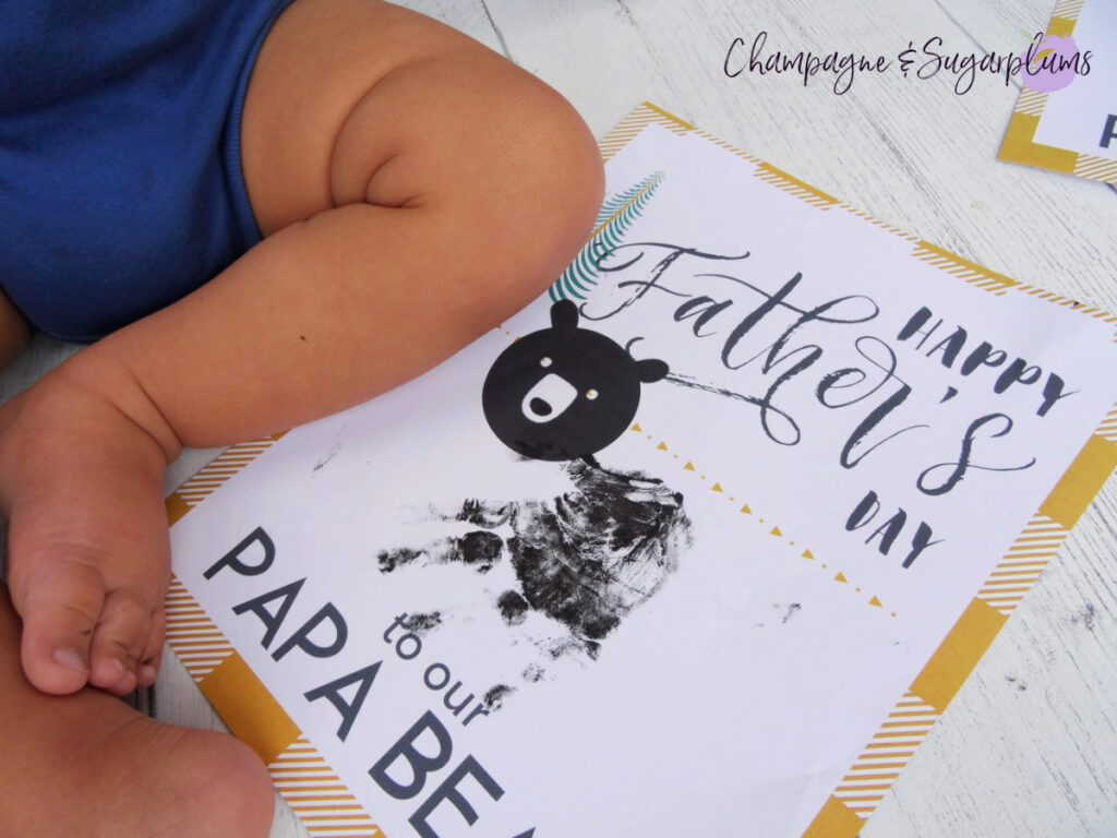 Papa Bear Handprint craft on a white background, with a toddler sitting beside it by Champagne and Sugarplums