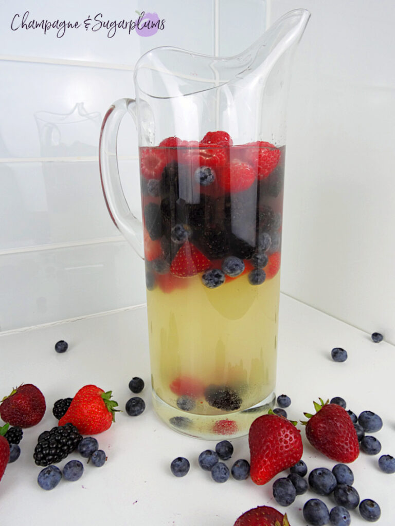 Red, Blue and Lychee Sangria in a pitcher, surrounded by berries on a white countertop by Champagne and Sugarplums