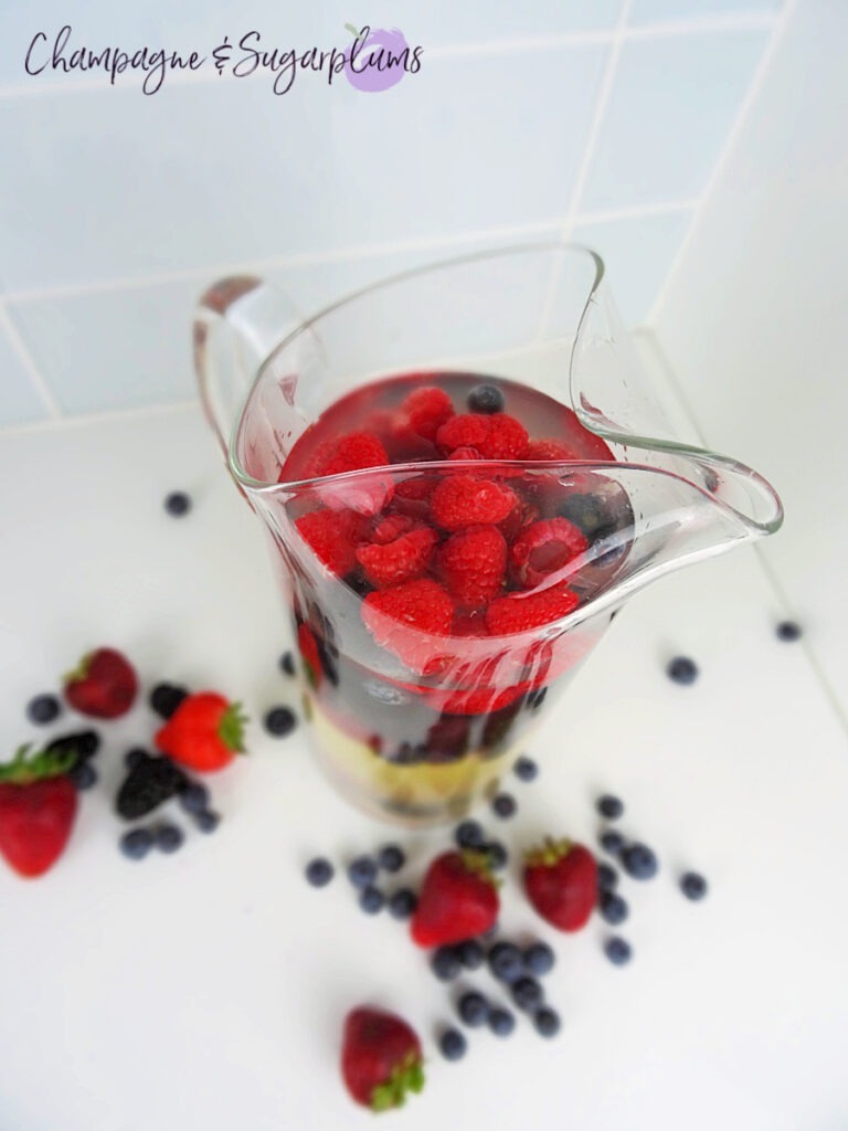 Red, Blue and Lychee Sangria in a pitcher, surrounded by berries on a white countertop by Champagne and Sugarplums