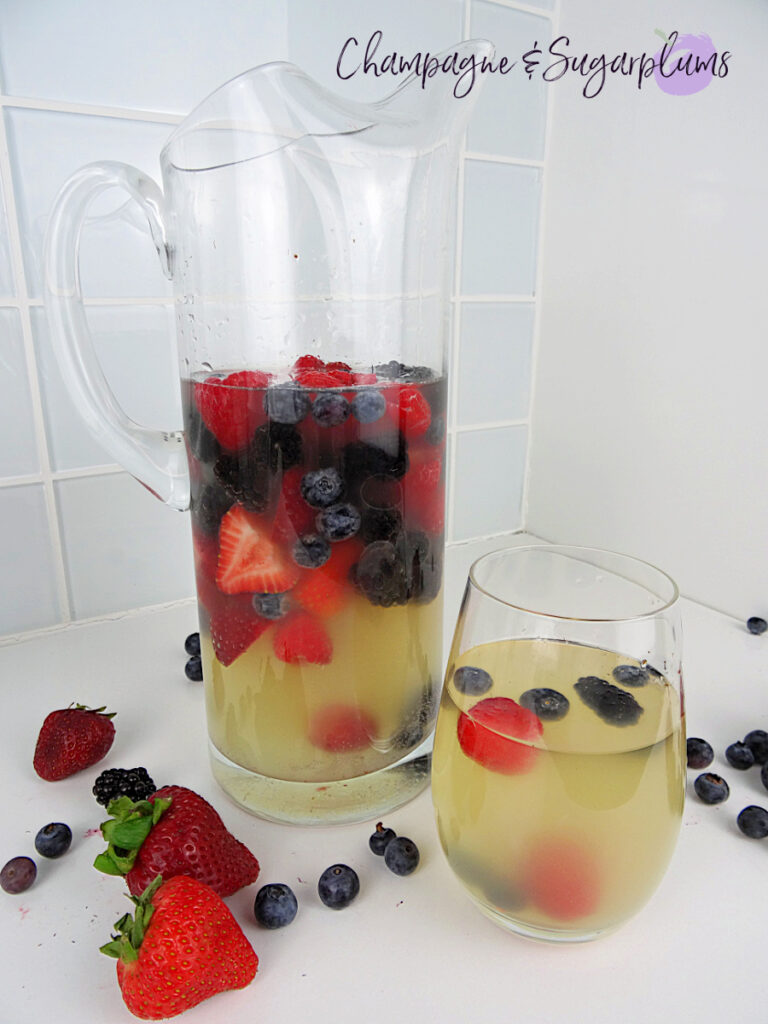 Red, Blue and Lychee Sangria in a glass and a pitcher, surrounded by berries on a white countertop by Champagne and Sugarplums
