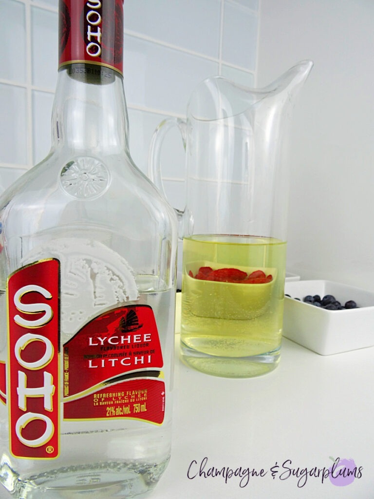 A pitcher beside a bottle of lychee liquor with white bowls of various berries on a white countertop by Champagne and Sugarplums