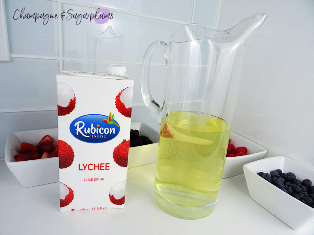 A pitcher beside a carton of lychee juice with white bowls of various berries on a white countertop by Champagne and Sugarplums