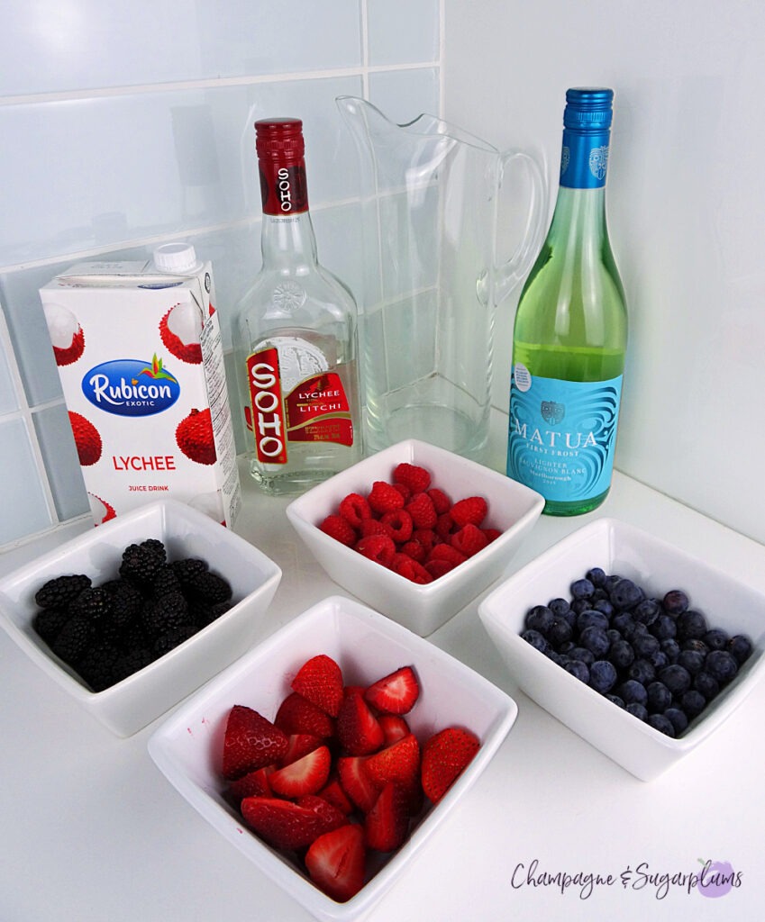 All the ingredients to make a Sangria in a pitcher, with berries in bowls on a white countertop by Champagne and Sugarplums