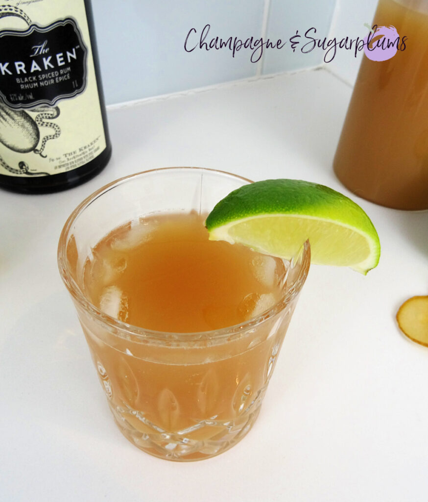 The Teacher's Pet Back to School Cocktail in a highball glass, garnished with a lime wedge on a white background with limes and a rum bottle by Champagne and Sugarplums